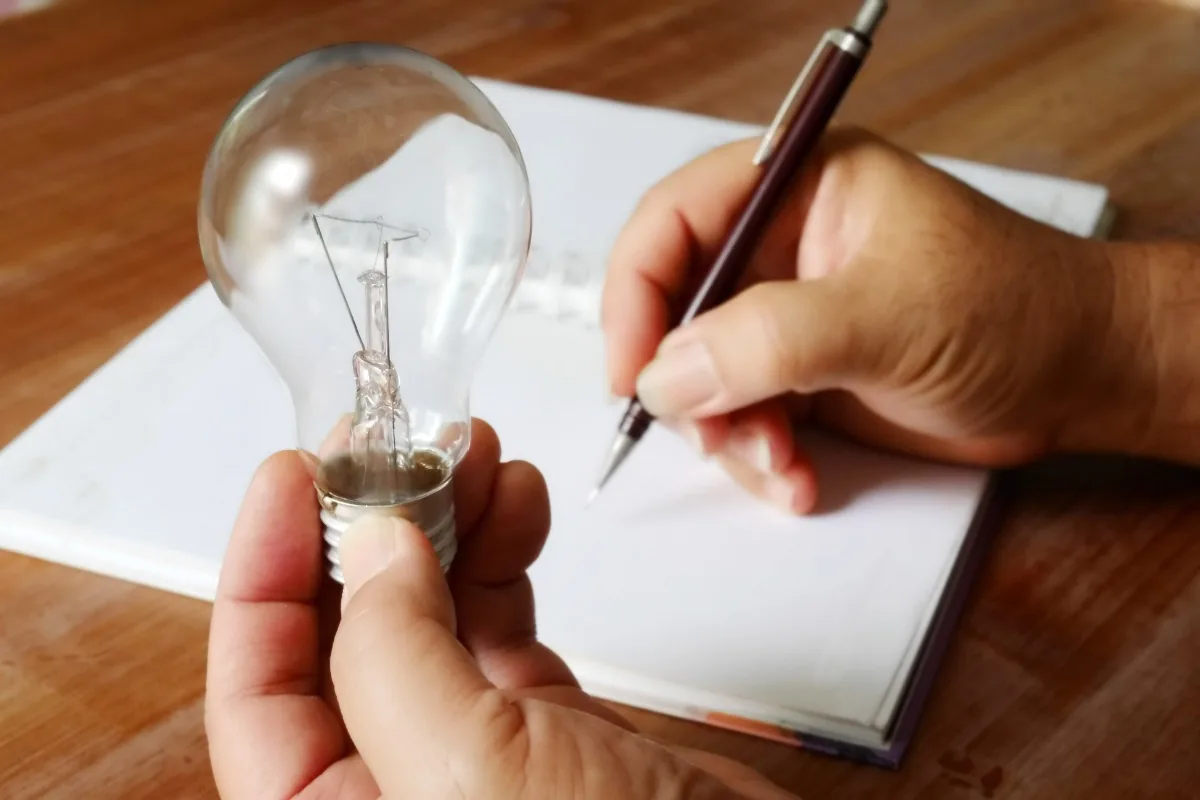 Hände halten eine Glühbirne und einen Stift über Unterlagen als Symbolbild für einen Energievertrag.